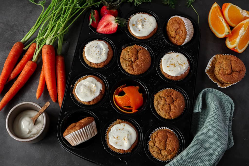Kalorienarme Karotten Muffins in einem Muffig Blech mit Frosting und Deko im Bildrand.