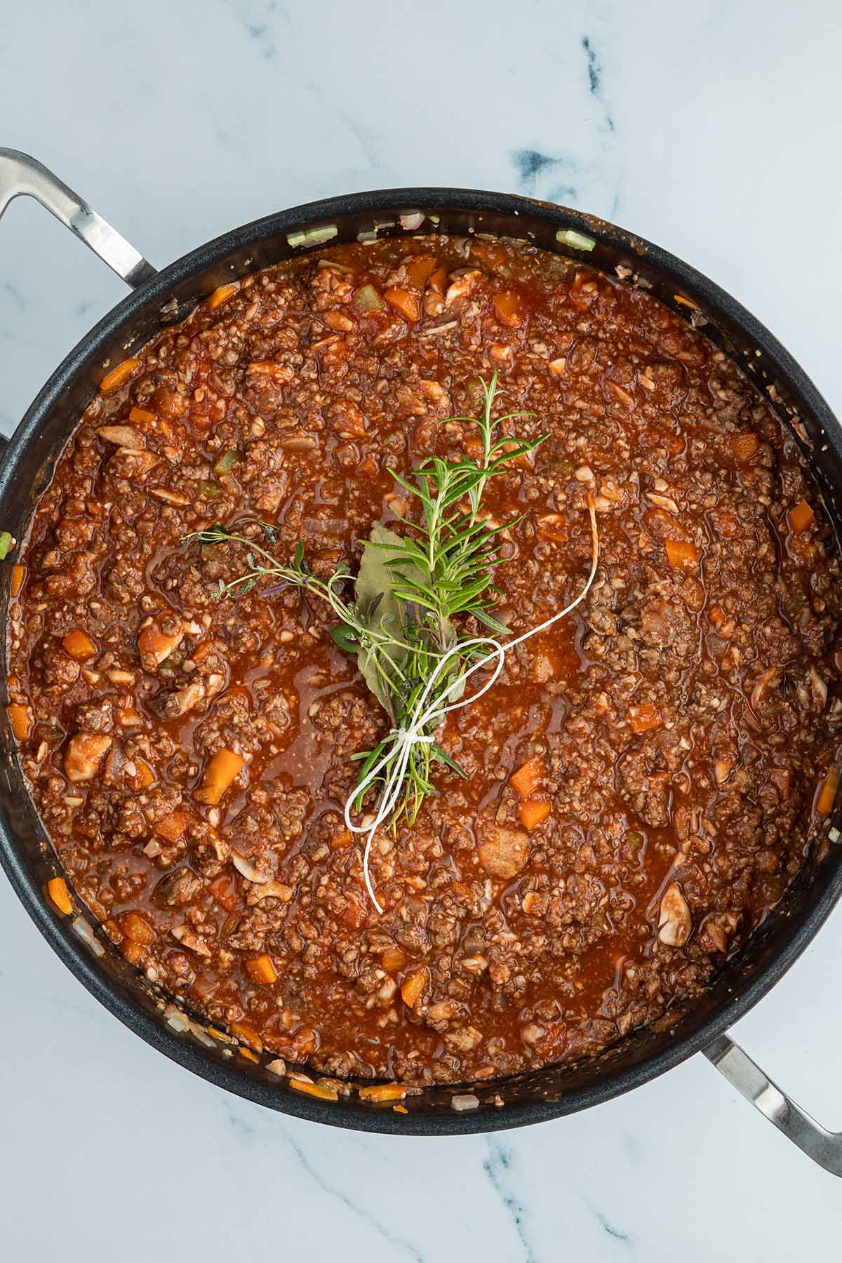 Vogelperspektive auf eine Schmorpfanne gef&uuml;llt mit einer Bolognese Sauce, auf deren Oberfl&auml;che ein Kr&auml;uterb&uuml;ndel liegt.