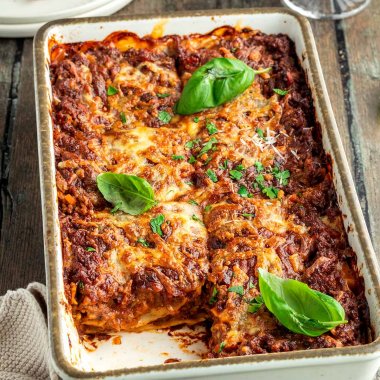 Eine herzhafte, mit frischen Kr&auml;utern servierte Lasagne in einer wei&szlig;en Auflaufform, ein Glas Rotwein und ein Teller mit einem St&uuml;ck der Lasagne im Hintergrund.