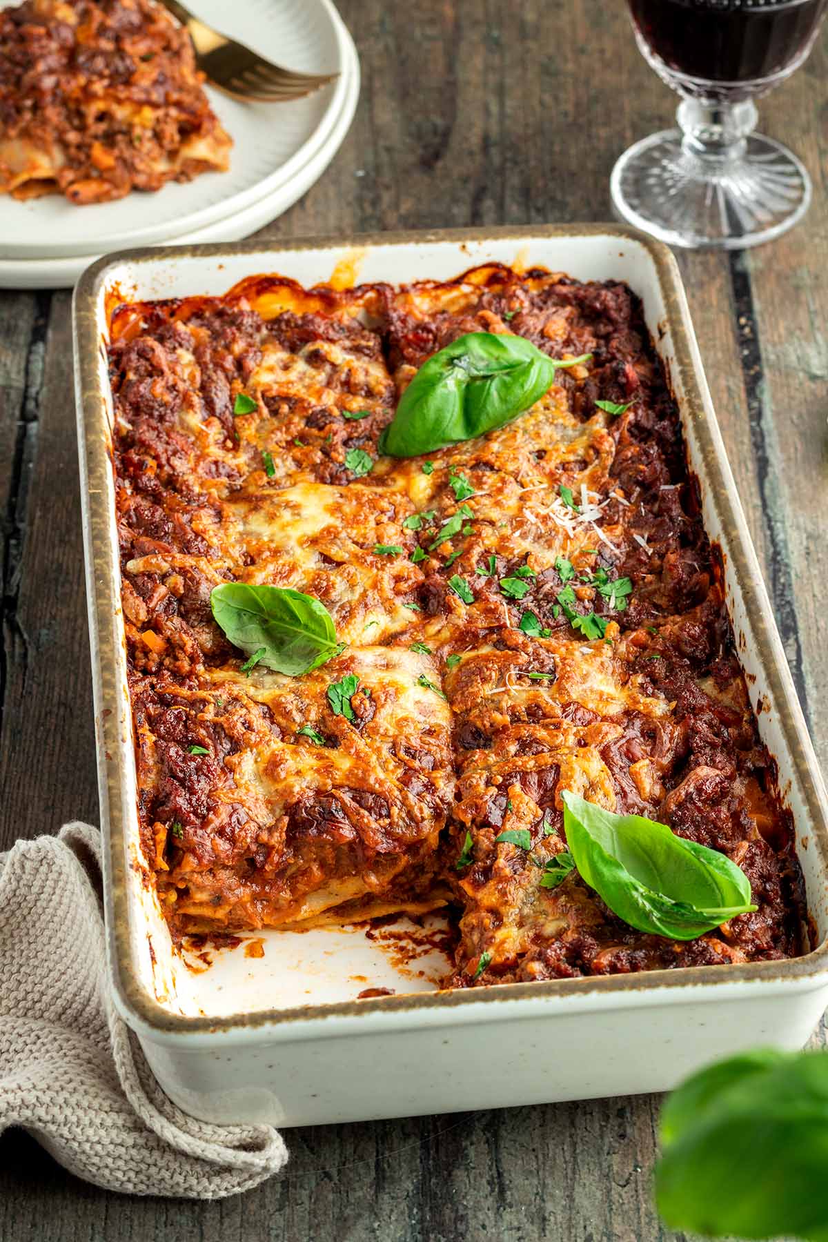Eine herzhafte, mit frischen Kr&auml;utern servierte Lasagne in einer wei&szlig;en Auflaufform, ein Glas Rotwein und ein Teller mit einem St&uuml;ck der Lasagne im Hintergrund.