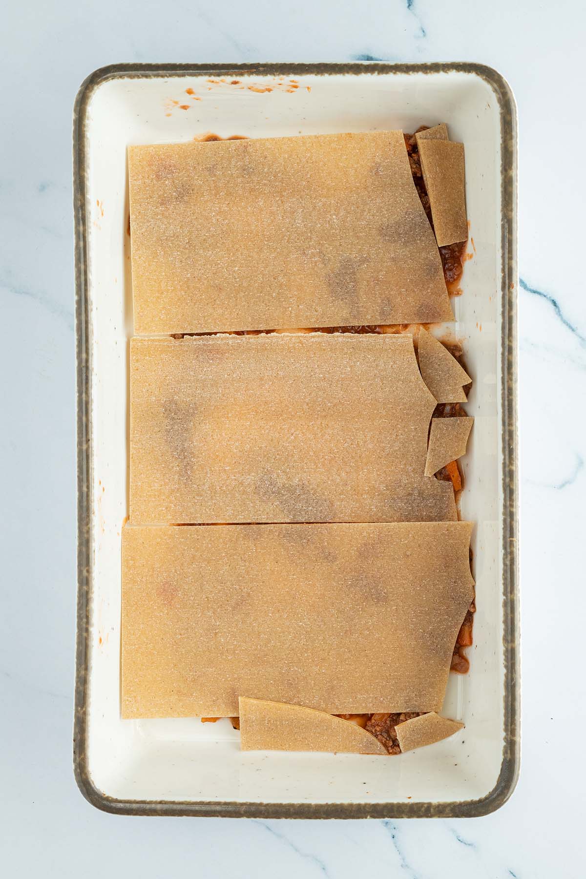 Vogelperspektive auf eine rechteckige Auflaufform, in der zurecht gebrochene Lasagne-Nudeln auf einer d&uuml;nnen Schicht von Sauce liegen.