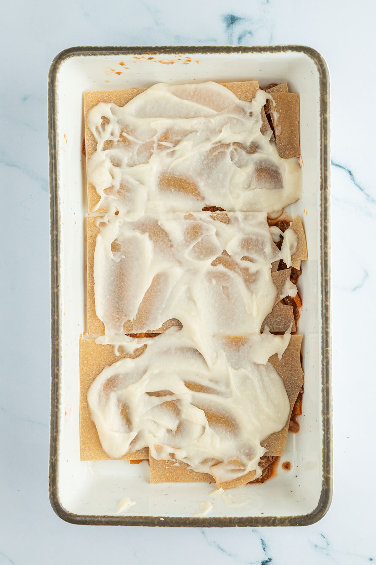 Vogelperspektive auf eine Auflaufform, in der zuerst etwas Fleischsauce, dann eine Schicht ungekochte Lasagne-Nudeln und darauf eine Schicht B&eacute;chamelsauce geschichtet wurde.