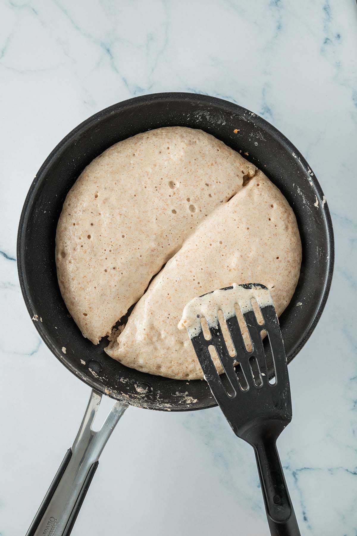 Ein mit einem Pfannenwender halbierter dicker, gro&szlig;er Pfannkuchen in einer Pfanne.