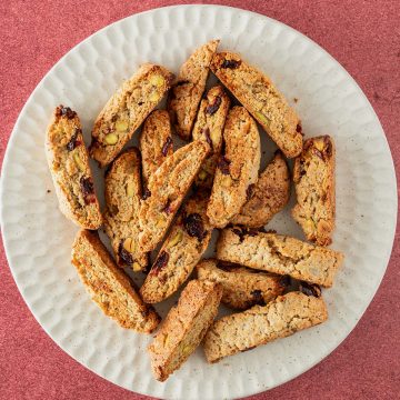Teller mit goldbraun gebackenen Kardamom-Pl&auml;tzchen mit Pistazien und Cranberries auf rotem Untergrund.