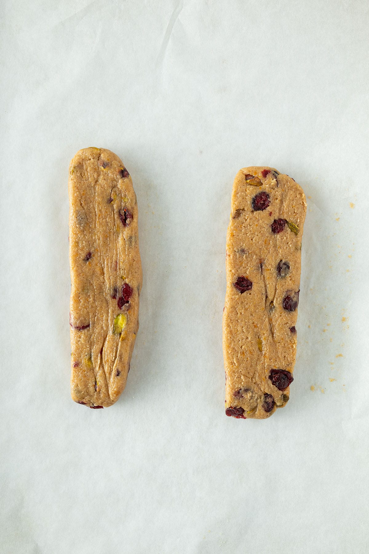 Zwei rohe Pl&auml;tzchen-Teigstr&auml;nge mit Pistazien und Cranberries auf Backpapier vorbereitet.