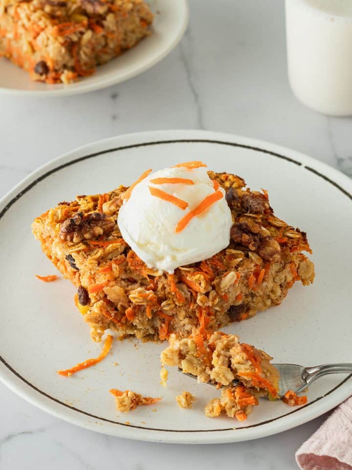 Ein St&uuml;ck Karotten-Baked-Oats auf einem Teller, serviert mit Joghurt. Ein zweiter Teller mit einem Glas Milch im Hintergrund.