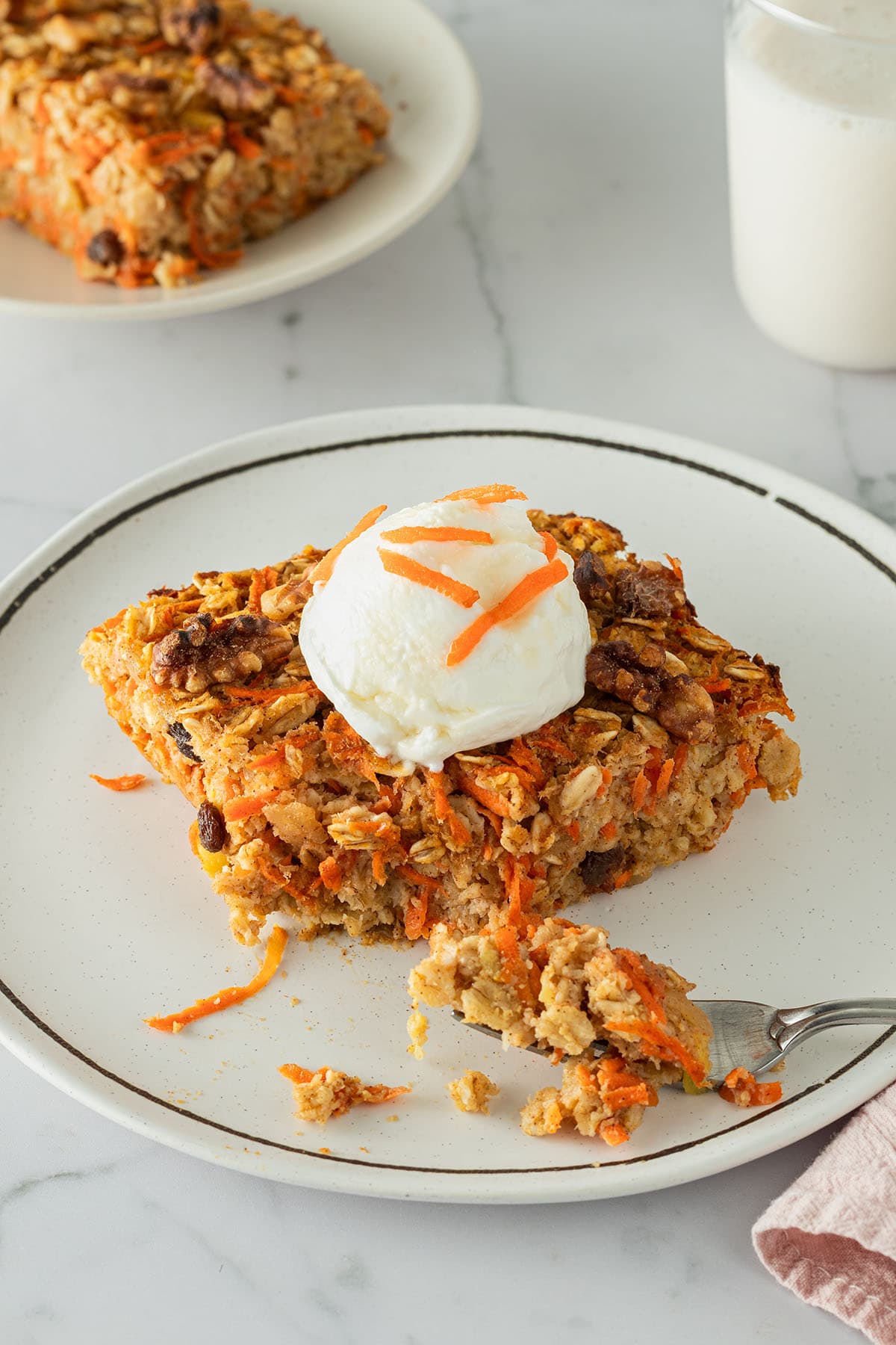 Ein St&uuml;ck Karotten-Baked-Oats auf einem Teller, serviert mit Joghurt. Ein zweiter Teller mit einem Glas Milch im Hintergrund.