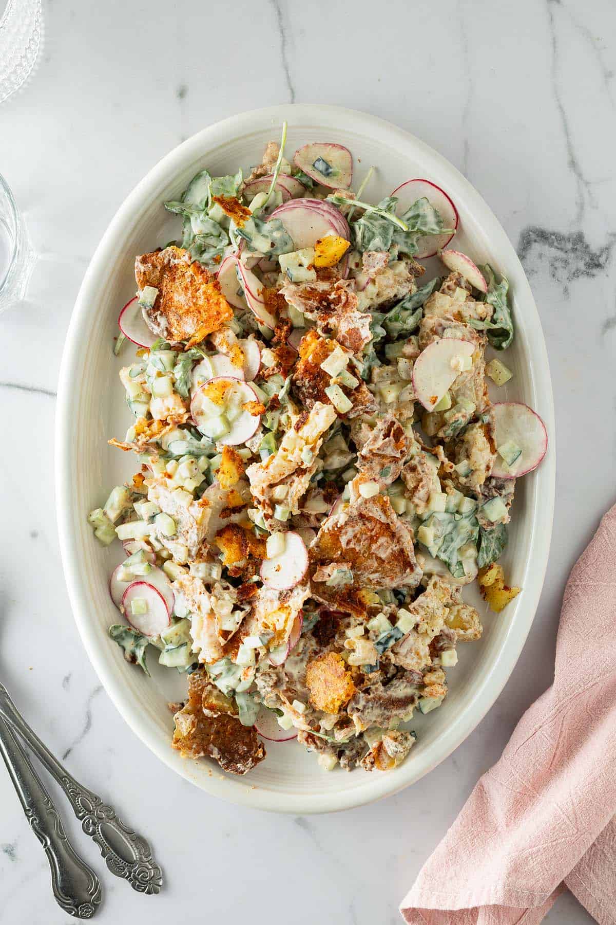 Knuspriger Kartoffelsalat mit ger&ouml;steten Kartoffeln, Gurken, Radieschen und einem cremigen Dressing, Gl&auml;ser un Besteck im Bildrand.