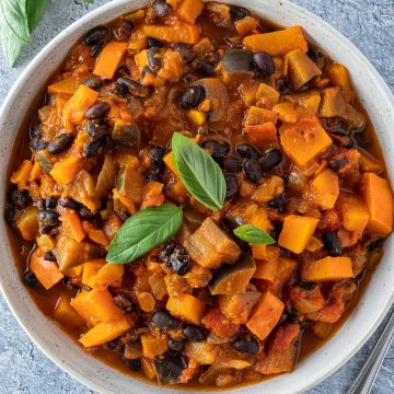 Ein Teller mit einem K&uuml;rbis-Bohnen-Curry mit Aubergine vor hellblauem Hintergrund.