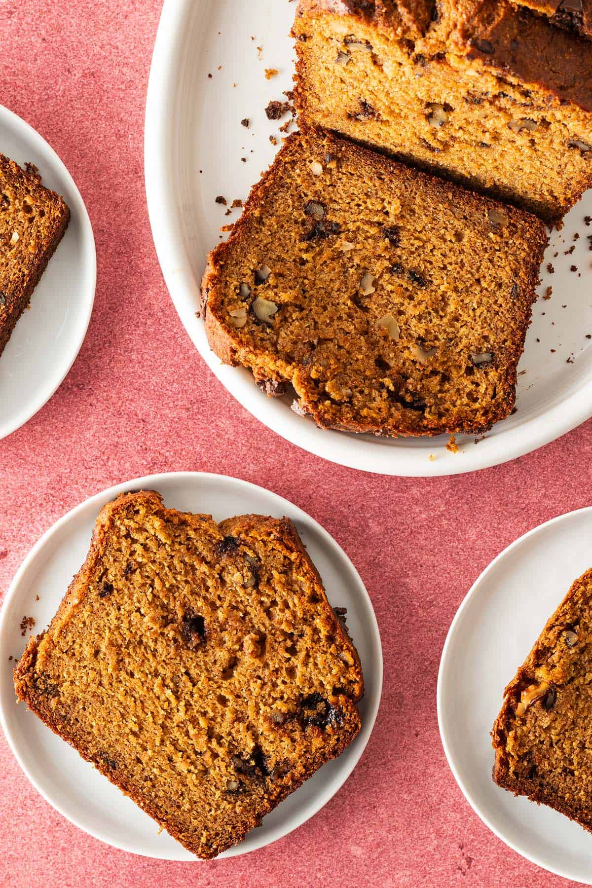 Mehrere Scheiben saftiges K&uuml;rbis-Bananenbrot mit Waln&uuml;ssen und Schokost&uuml;ckchen auf wei&szlig;en Tellern, angerichtet auf rosa Untergrund.