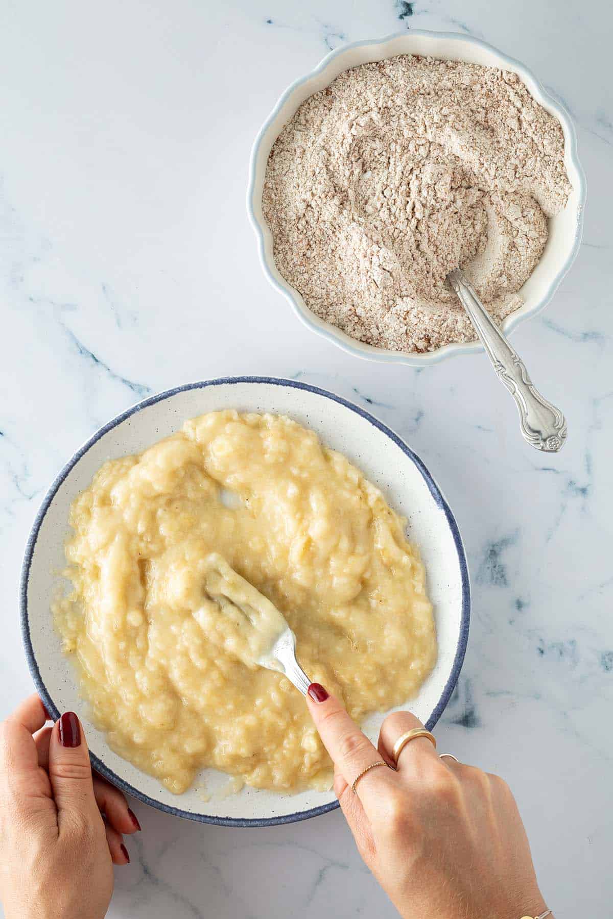 Zwei Sch&uuml;sseln mit Zutaten f&uuml;r Bananenbrot: eine Sch&uuml;ssel mit zerdr&uuml;ckten Bananen, die mit einer Gabel bearbeitet werden, daneben eine Sch&uuml;ssel mit Mehlmischung und L&ouml;ffel.