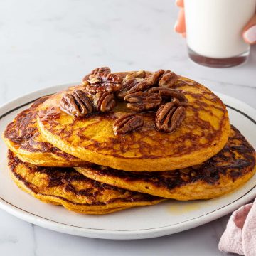 Ein Stapel K&uuml;rbis Pancakes auf einem Teller mit N&uuml;ssen und Ahornsirup. Eine Hand h&auml;lt ein Glas Milch im Hintergrund.