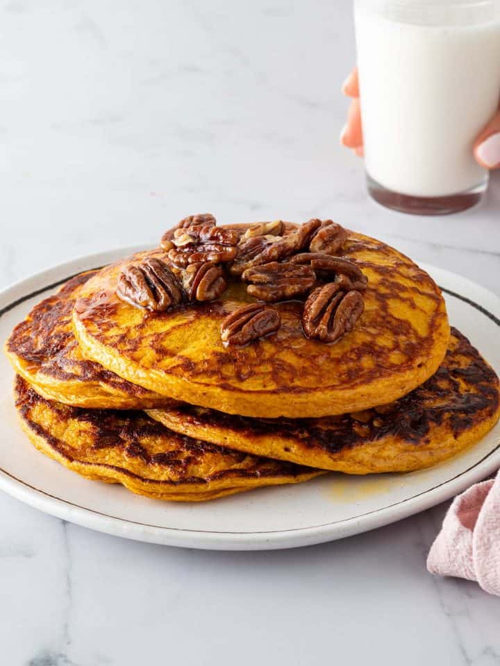 Ein Stapel K&uuml;rbis Pancakes auf einem Teller mit N&uuml;ssen und Ahornsirup. Eine Hand h&auml;lt ein Glas Milch im Hintergrund.