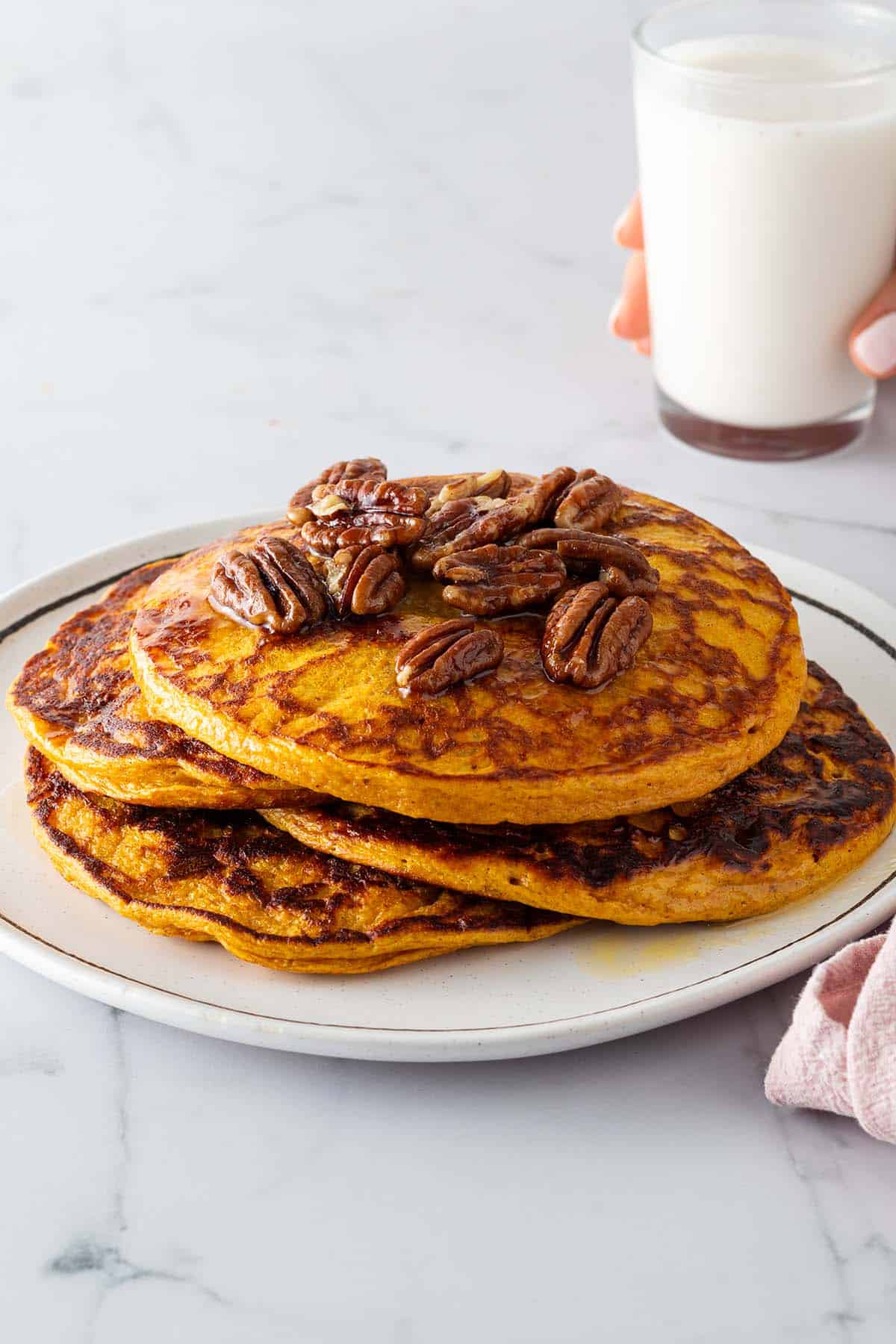 Ein Stapel K&uuml;rbis Pancakes auf einem Teller mit N&uuml;ssen und Ahornsirup. Eine Hand h&auml;lt ein Glas Milch im Hintergrund.
