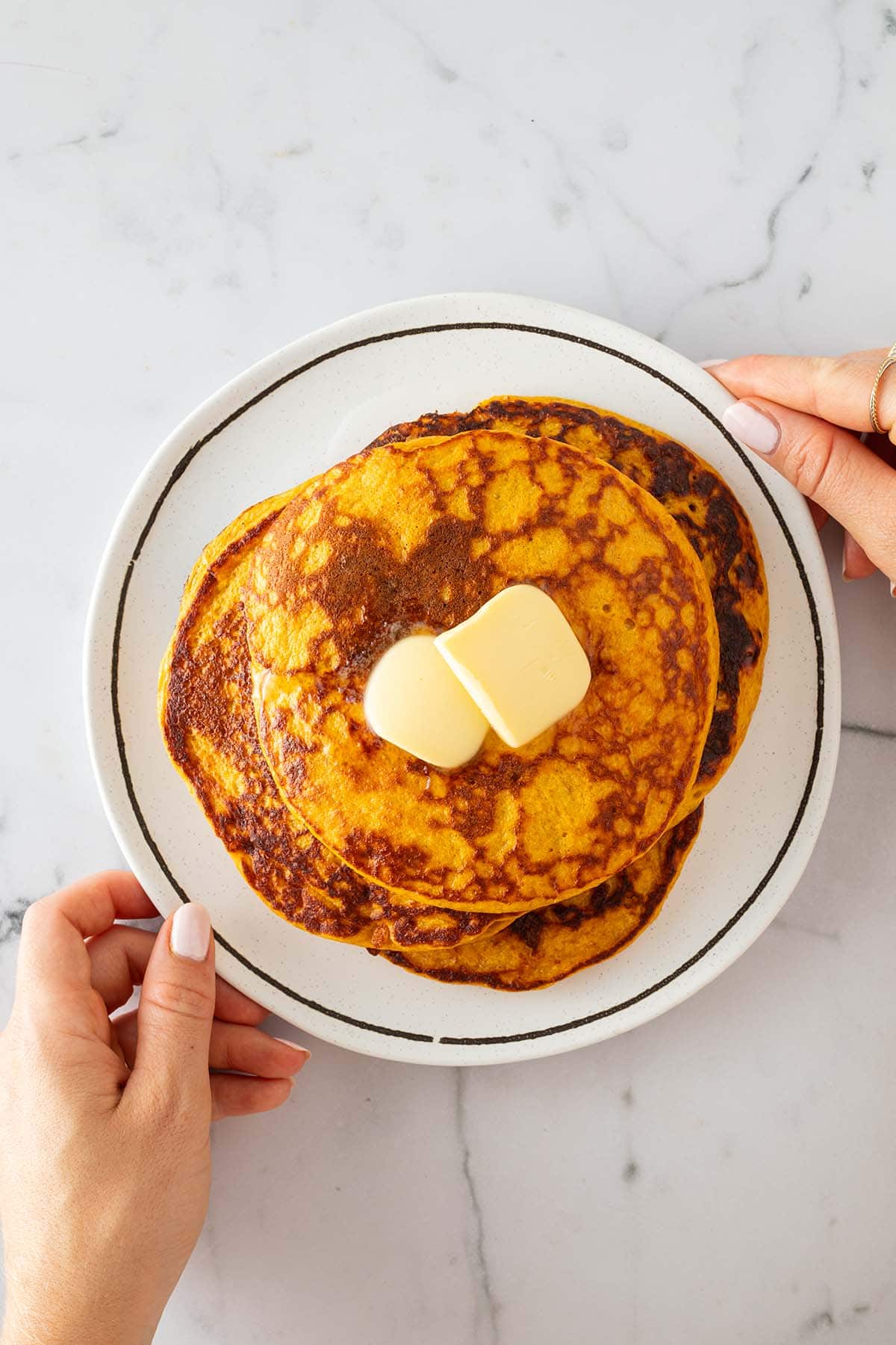Eine Person h&auml;lt ein Teller mit mehreren K&uuml;rbis Pancakes in der Hand mit zwei St&uuml;cke Butter auf dem obersten Pancake.