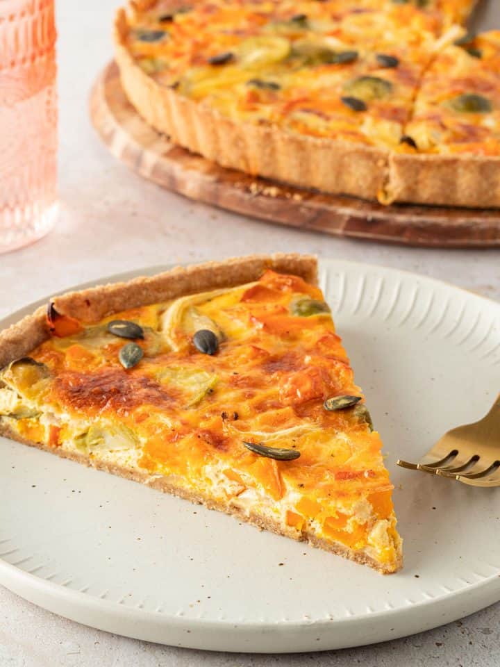 Ein St&uuml;ck K&uuml;rbis Quiche mit Rosenkohl und K&uuml;rbiskernen, die restliche Quiche im Hintergrund.