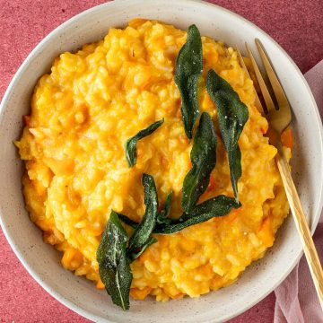 Cremige, oranges K&uuml;rbis Risotto mit knusprigen Salbei auf einem roten Untergrund, Deko im Bildrand.