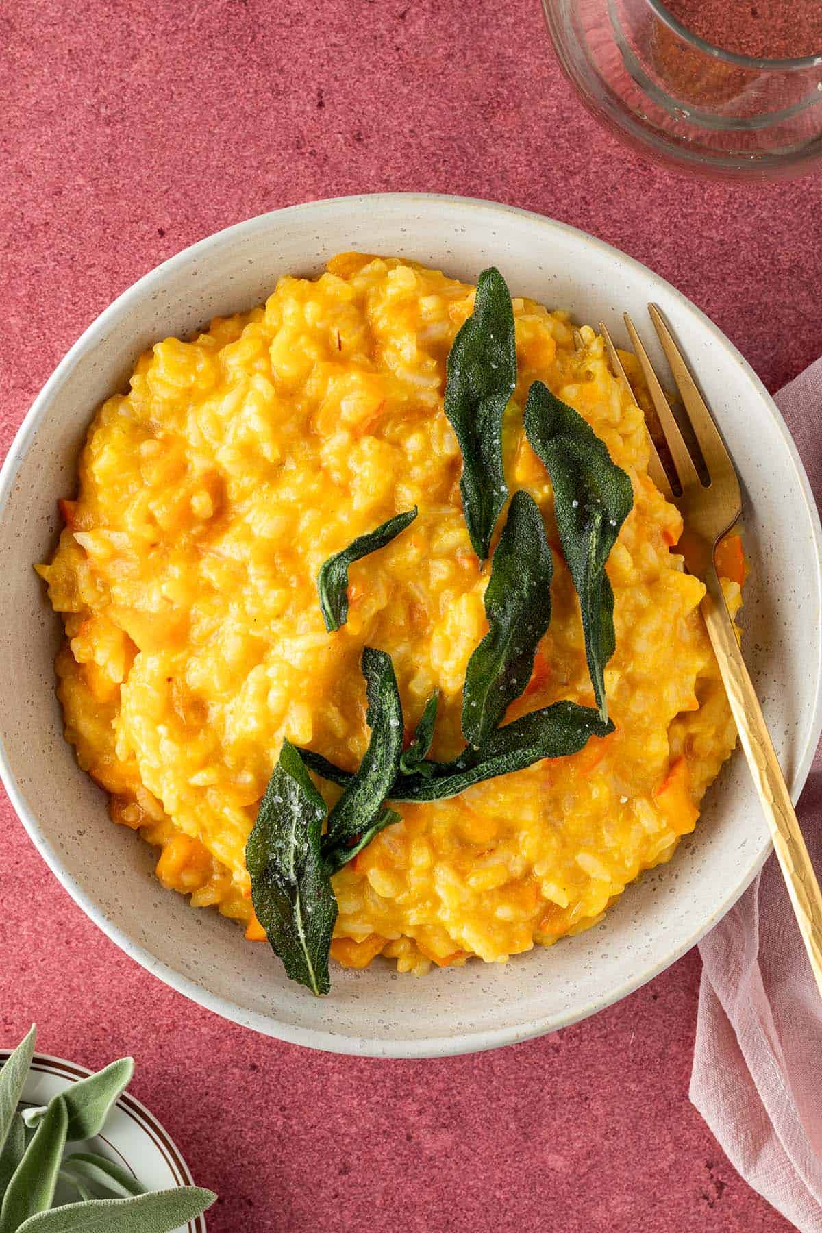Cremige, oranges K&uuml;rbis Risotto mit knusprigen Salbei auf einem roten Untergrund, Deko im Bildrand.