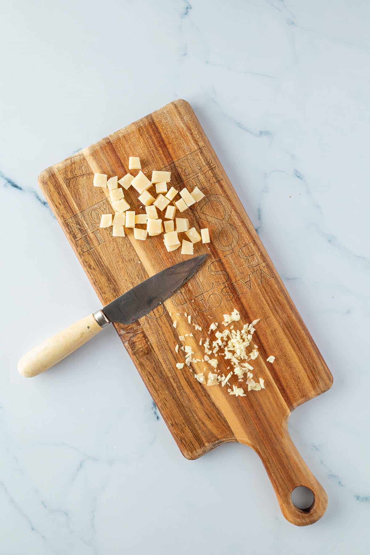 Fein gew&uuml;rfelter Knoblauch und kleine Parmesanw&uuml;fel auf einem Holzbrett mit einem kleinen K&uuml;chenmesser daneben.