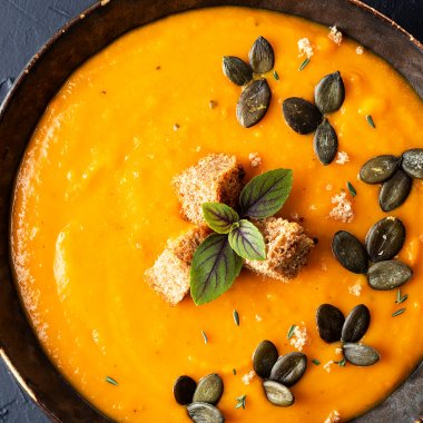 Eine Sch&uuml;ssel mit K&uuml;rbissuppe serviert mit ger&ouml;steten Brotw&uuml;rfeln und K&uuml;rbiskernen auf einem dunkelblauen Hintergrund.