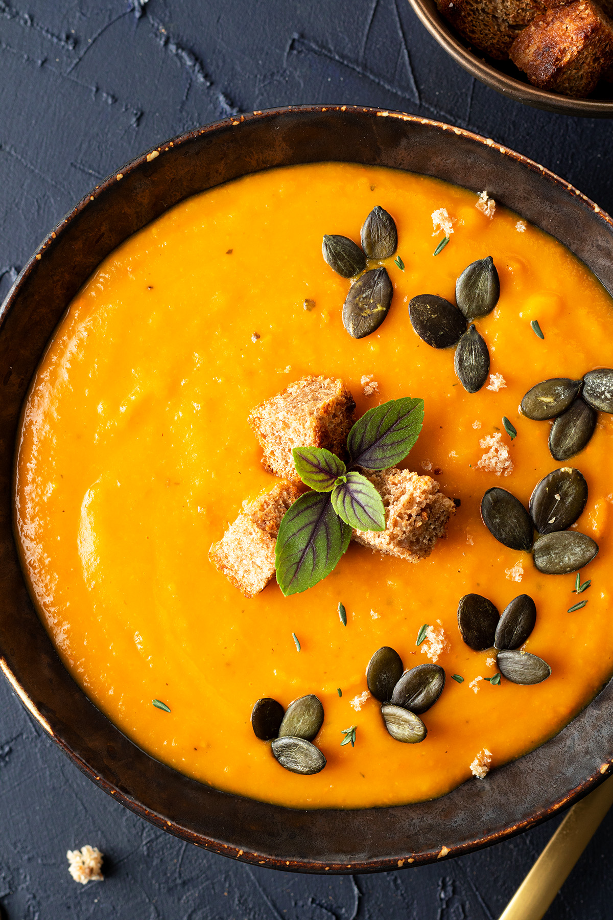 Eine Sch&uuml;ssel mit K&uuml;rbissuppe serviert mit ger&ouml;steten Brotw&uuml;rfeln und K&uuml;rbiskernen auf einem dunkelblauen Hintergrund.