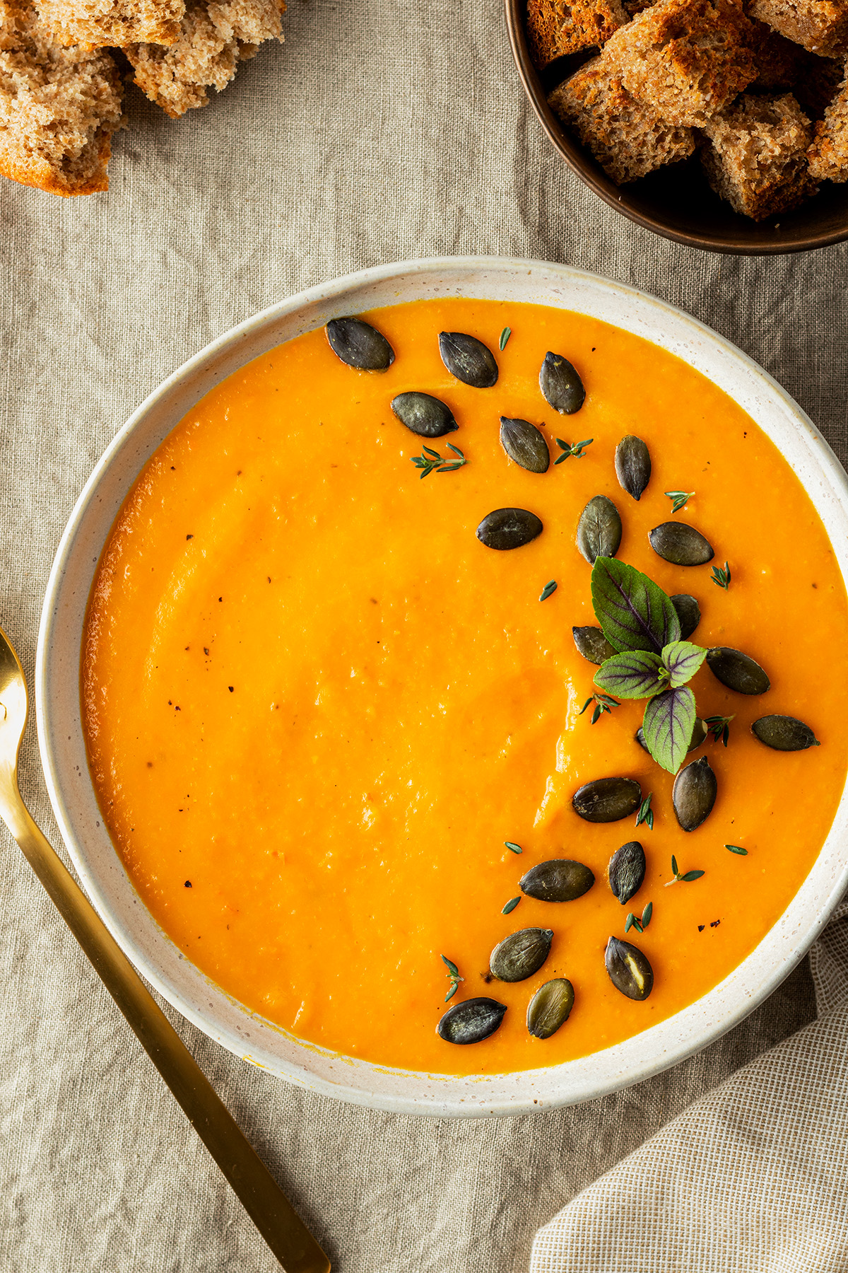 Ein Teller K&uuml;rbissuppe mit K&uuml;rbiskernen verziert auf einer beigen Tischdecke. Daneben eine Sch&uuml;ssel mit ger&ouml;steten Brotw&uuml;rfeln.