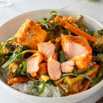 Eine Sch&uuml;ssel mit Thai Lachs Curry mit saftigen Lachsw&uuml;rfeln, Karotten und Brokkoli serviert auf Reis mit frischem Basilikum. Im Hintergrund ein Glas Wasser, eine Limette und Esst&auml;bchen im Bildrand.