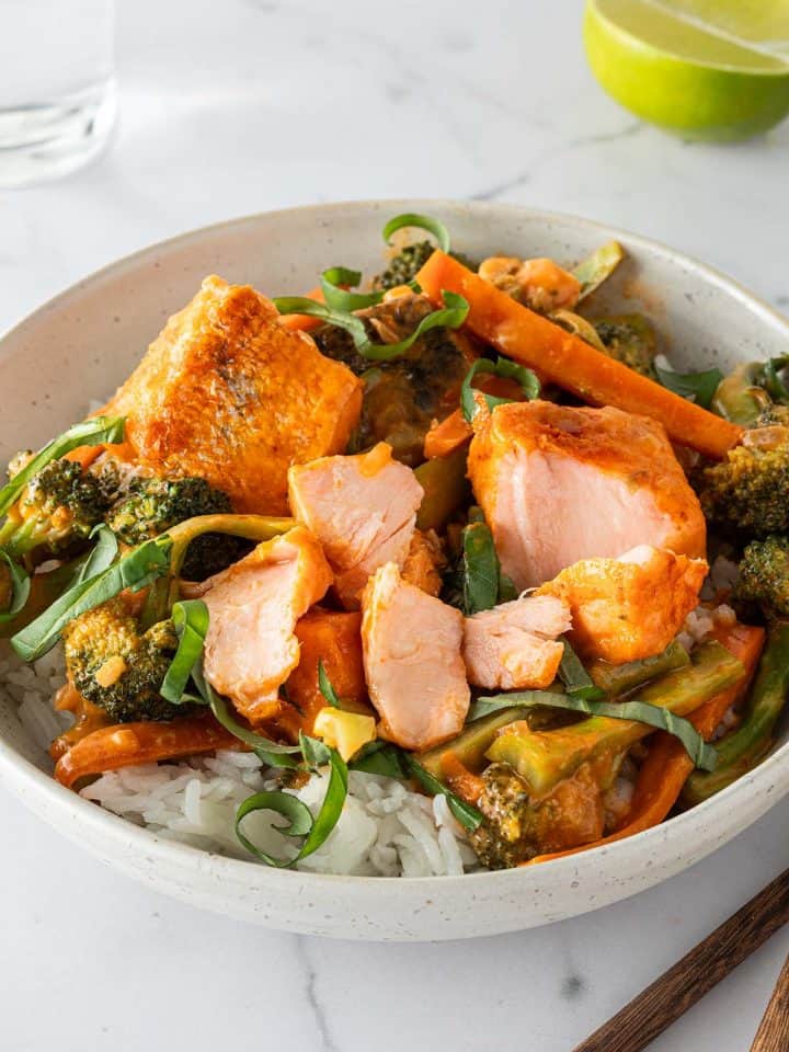 Eine Sch&uuml;ssel mit Thai Lachs Curry mit saftigen Lachsw&uuml;rfeln, Karotten und Brokkoli serviert auf Reis mit frischem Basilikum. Im Hintergrund ein Glas Wasser, eine Limette und Esst&auml;bchen im Bildrand.