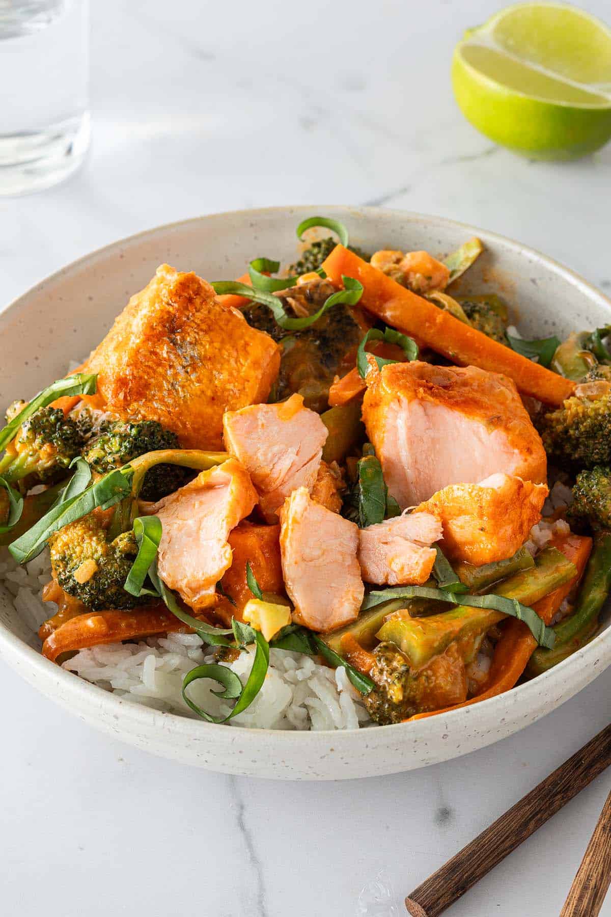 Eine Sch&uuml;ssel mit Thai Lachs Curry mit saftigen Lachsw&uuml;rfeln, Karotten und Brokkoli serviert auf Reis mit frischem Basilikum. Im Hintergrund ein Glas Wasser, eine Limette und Esst&auml;bchen im Bildrand.