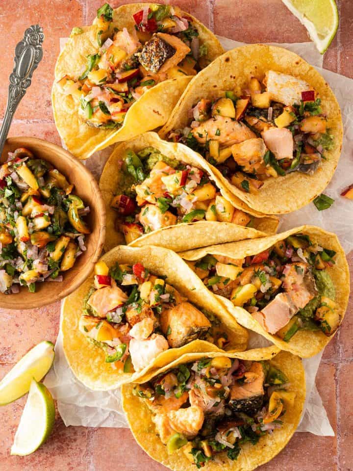 Mehrere Tacos mit Lachs St&uuml;ckchen, Avocadocreme und Pfirsich Salsa auf einem roten Fliesenuntergrund, Limettenspalten und eine Sch&uuml;ssel Pfirsich Salsa im Bildrand.