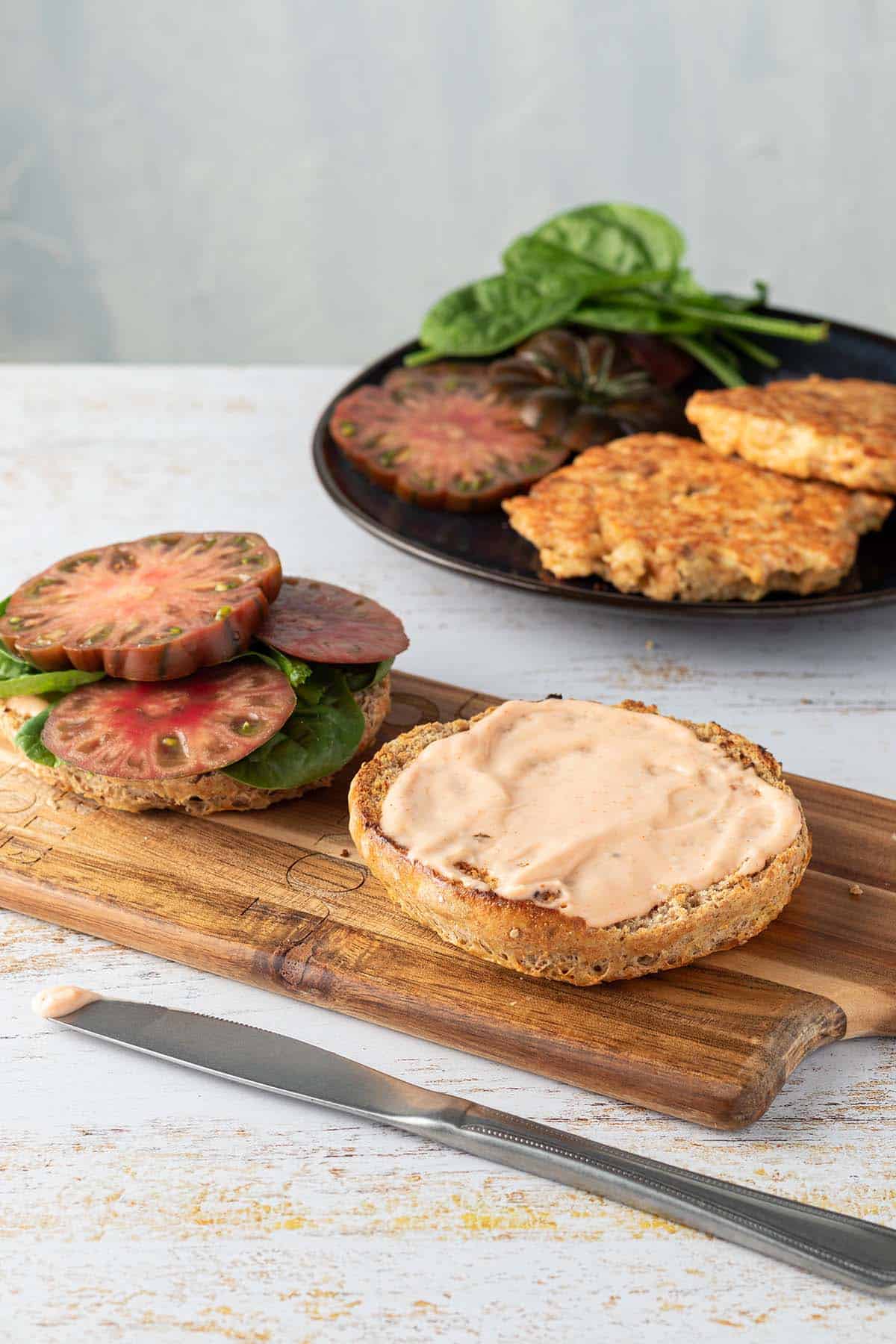 Ein Burger Brötchen wird mit Sriracha Mayo, Spinat und Tomaten belegt, im Hintergrund ein Teller mit goldbraunen Lachsfilets, Tomaten-Scheiben und Spiantblättern.