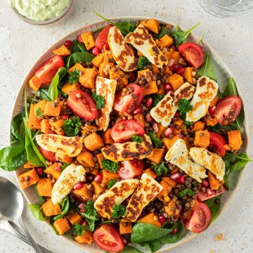 Linsensalat mit frischem Spinat, ger&ouml;steten S&uuml;&szlig;kartoffeln, Tomaten, gebratenem Halloumi und Granatapfelkernen auf einem gro&szlig;en Servierteller mit einer Sch&uuml;ssel Avocado-Dressing daneben.