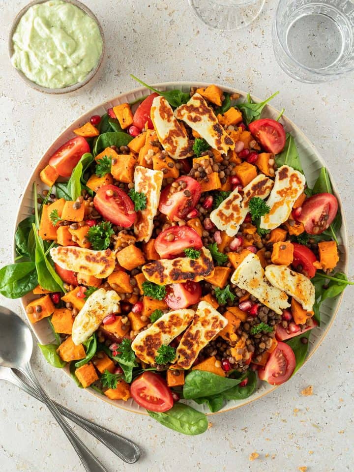 Linsensalat mit frischem Spinat, ger&ouml;steten S&uuml;&szlig;kartoffeln, Tomaten, gebratenem Halloumi und Granatapfelkernen auf einem gro&szlig;en Servierteller mit einer Sch&uuml;ssel Avocado-Dressing daneben.