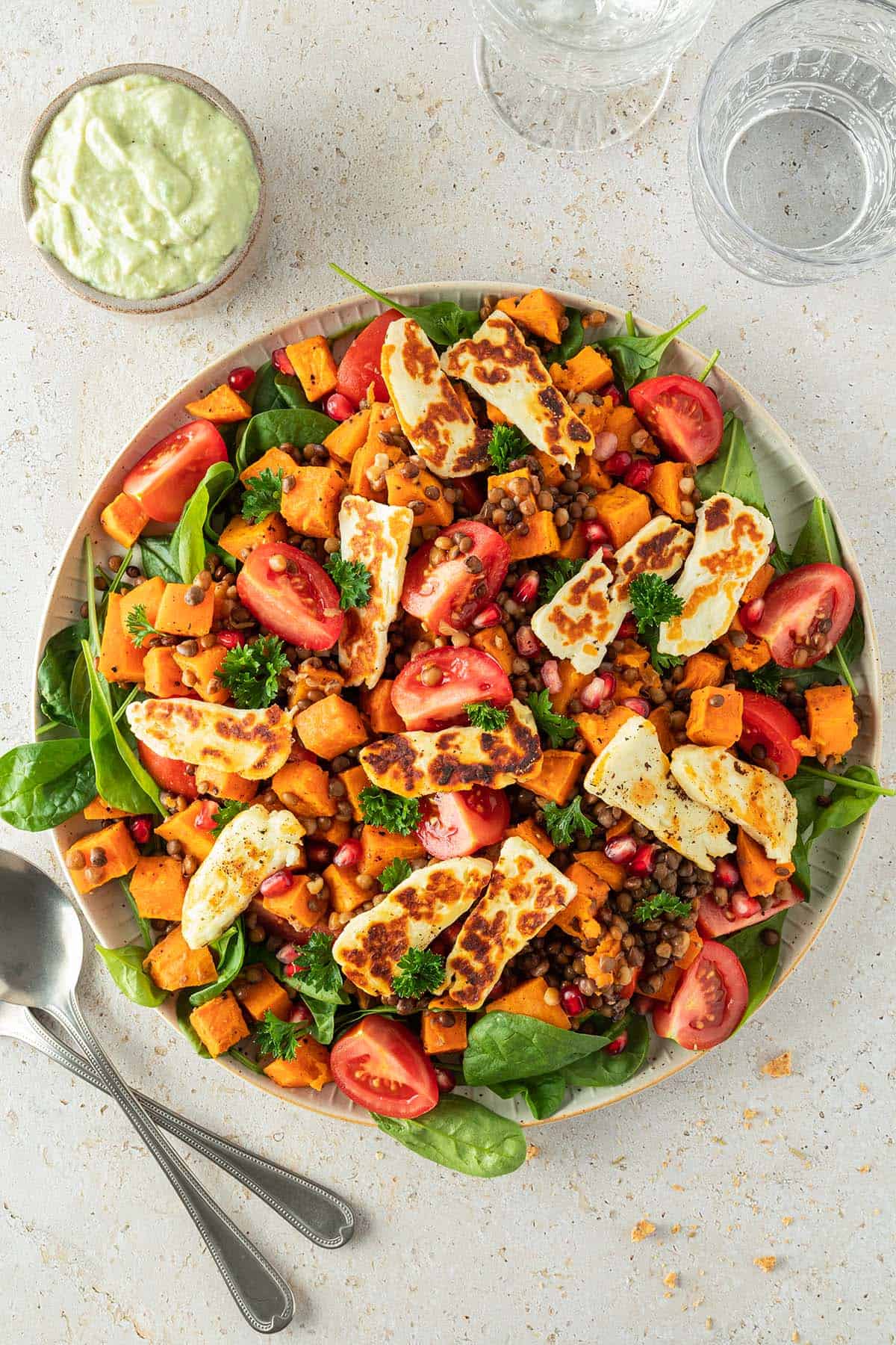 Linsensalat mit frischem Spinat, ger&ouml;steten S&uuml;&szlig;kartoffeln, Tomaten, gebratenem Halloumi und Granatapfelkernen auf einem gro&szlig;en Servierteller mit einer Sch&uuml;ssel Avocado-Dressing daneben.