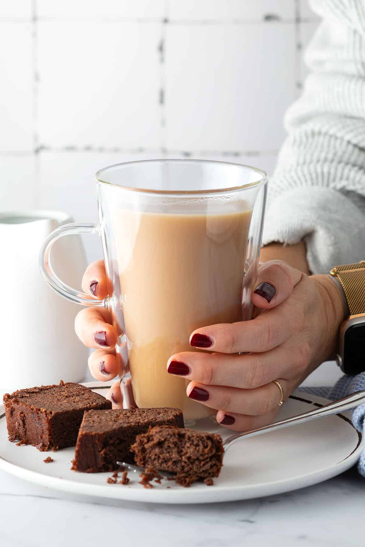 Eine Person h&auml;lt eine Tasse London Fog in den H&auml;nden, ein St&uuml;ck Schokokuchen und ein wei&szlig;es K&auml;nnchen sind im Bild zu sehen.