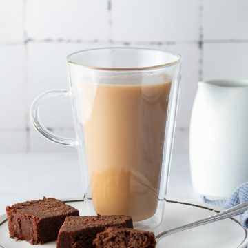 Eine Glastasse London Fog auf einem Teller mit einem St&uuml;ck Schokokuchen, eine Milchkanne im Hintergrund.
