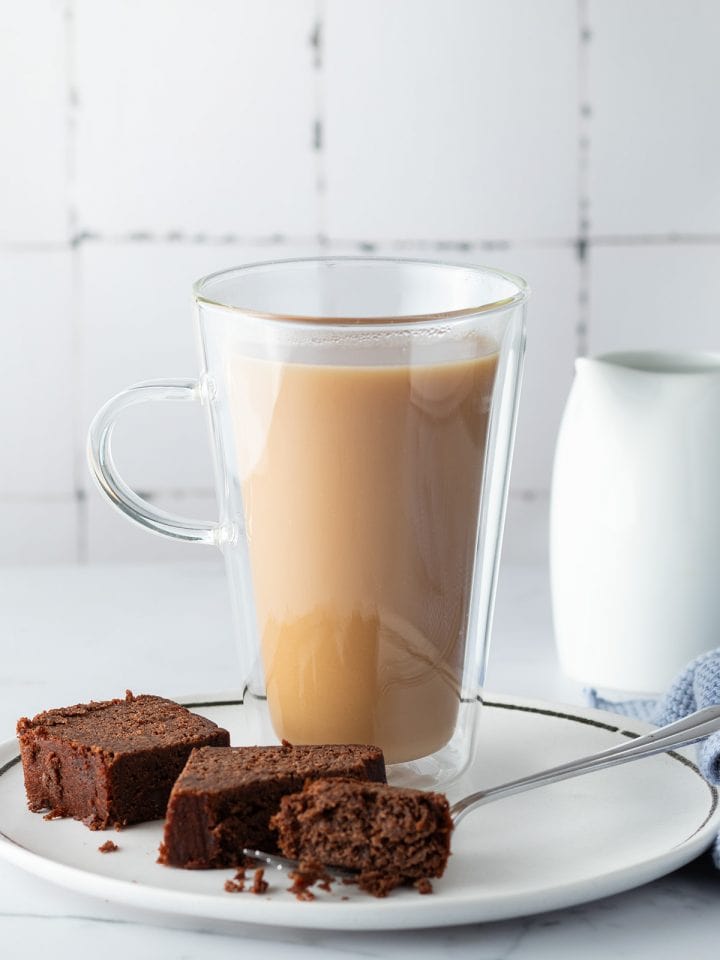 Eine Glastasse London Fog auf einem Teller mit einem St&uuml;ck Schokokuchen, eine Milchkanne im Hintergrund.