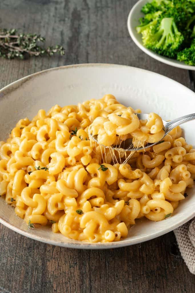 Ein Teller Mac and Cheese mit einem L&ouml;ffel, im Hintergrund ein Teller Brokkoli.