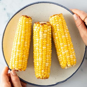 Drei im Airfryer ger&ouml;stete Maiskolben auf einem Teller, von zwei H&auml;nden gehalten.