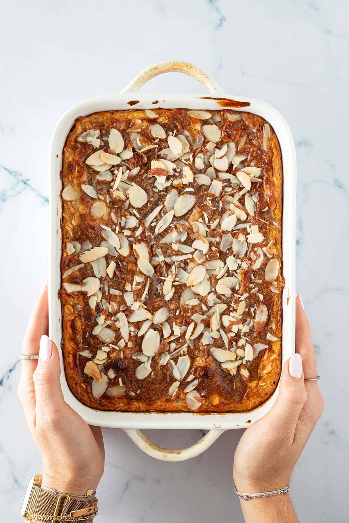 Frisch gebackene Mandel-Croissant Baked Oats direkt nach dem Backen in einer wei&szlig;en Auflaufform, von zwei H&auml;nden gehalten.