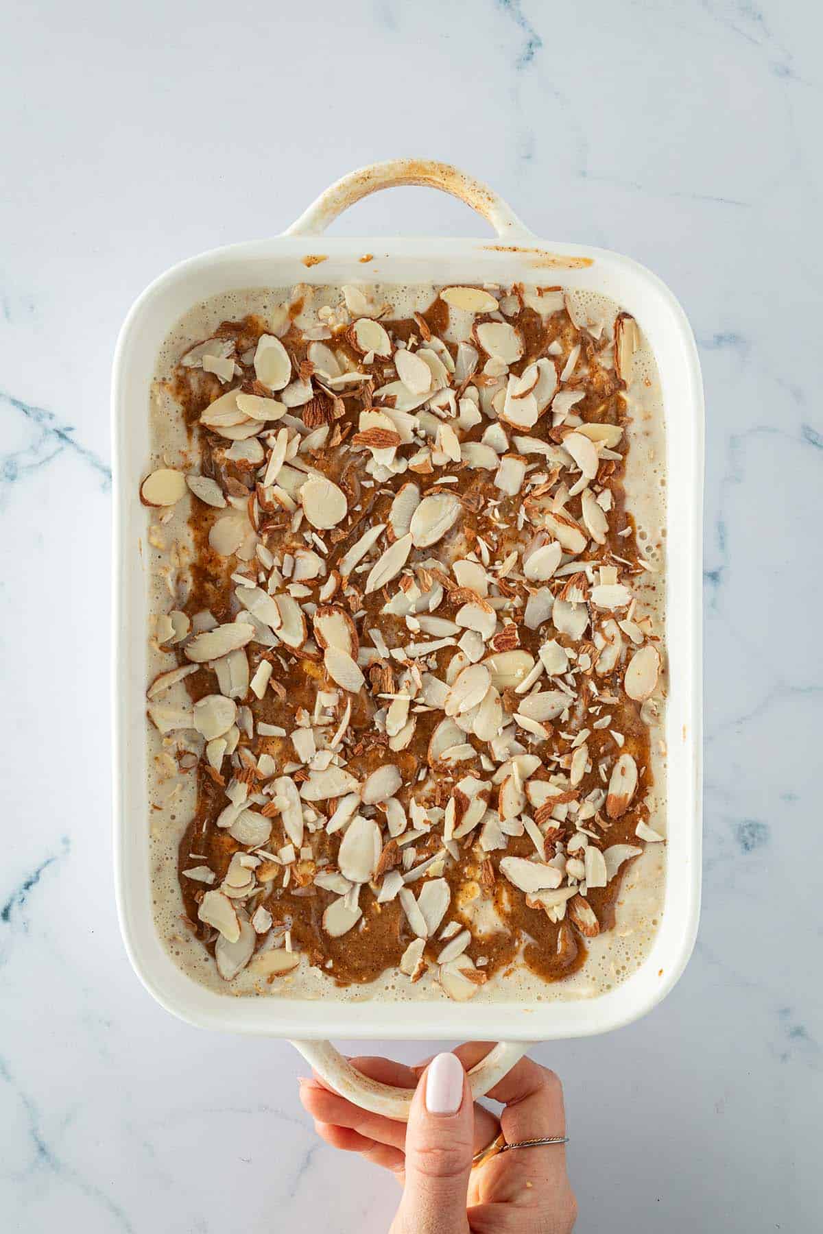 Ungebackene Mandel-Croissant Baked Oats-Masse in einer wei&szlig;en Auflaufform, mit Mandelbl&auml;ttchen und einer Mandelmusmischung on top.