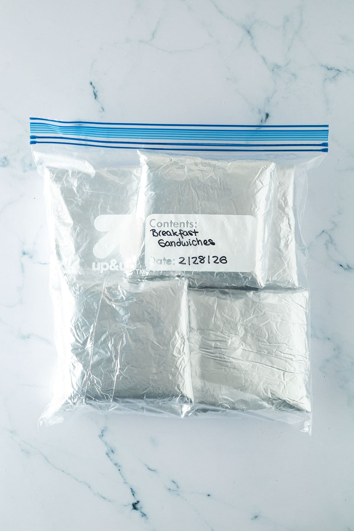 Sechs in Folie gewickelte Fr&uuml;hst&uuml;ckssandwiches in einem beschrifteten Gefrierbeutel zum Meal Prep eingefroren.
