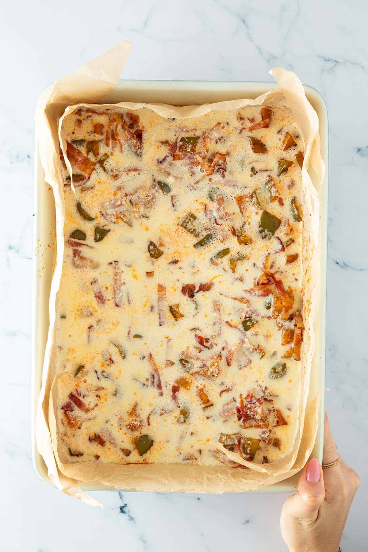 Auflaufform mit Backpapier ausgelegt und gef&uuml;llt mit roher Eier-H&uuml;ttenk&auml;se-Mischung, Kartoffeln, Paprika und Speckstreifen.