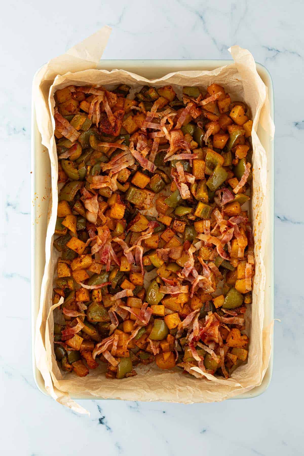 Ger&ouml;stete Kartoffelw&uuml;rfel, gr&uuml;ne Paprika und knusprige Speckstreifen in einer mit Backpapier ausgelegten Auflaufform.