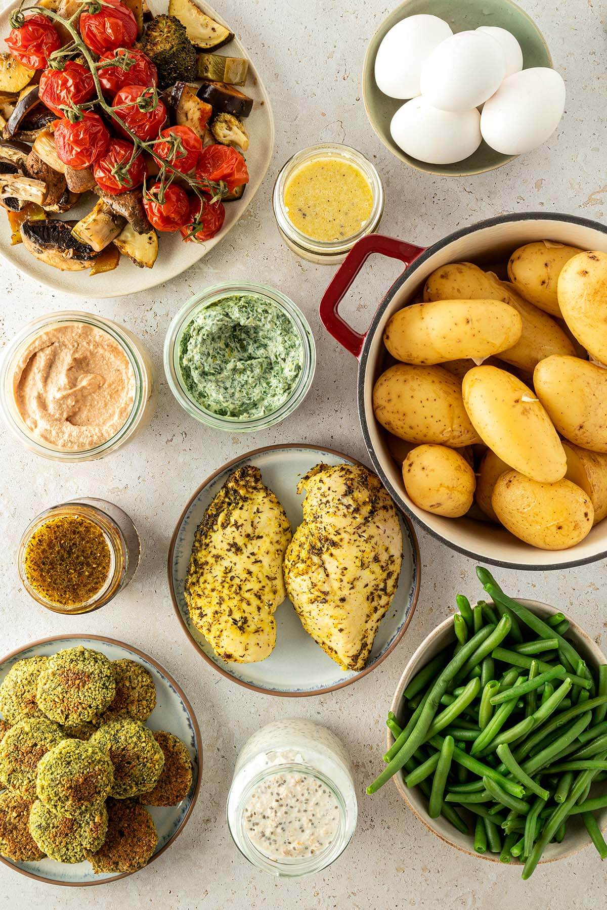 Ofengem&uuml;se, Eier, Gl&auml;ser mit Dressings und Saucen, Falafel, mariniertes H&auml;hnchen, gekochte Bohnen und Kartoffeln auf einem beigen Untergrund.