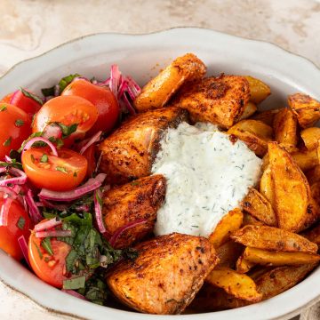 Mediterrane Lachs Bowl mit w&uuml;rzigen Lachsw&uuml;rfeln, knusprigen Kartoffelspalten, Tomatensalat und cremigem Tzatziki auf einem hellen Steinuntergrund.