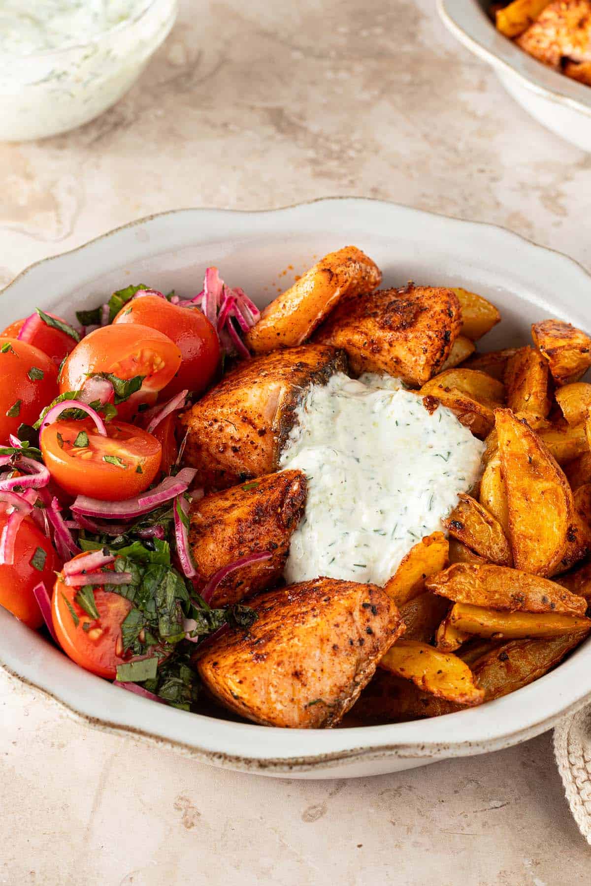 Mediterrane Lachs Bowl mit w&uuml;rzigen Lachsw&uuml;rfeln, knusprigen Kartoffelspalten, Tomatensalat und cremigem Tzatziki auf einem hellen Steinuntergrund.
