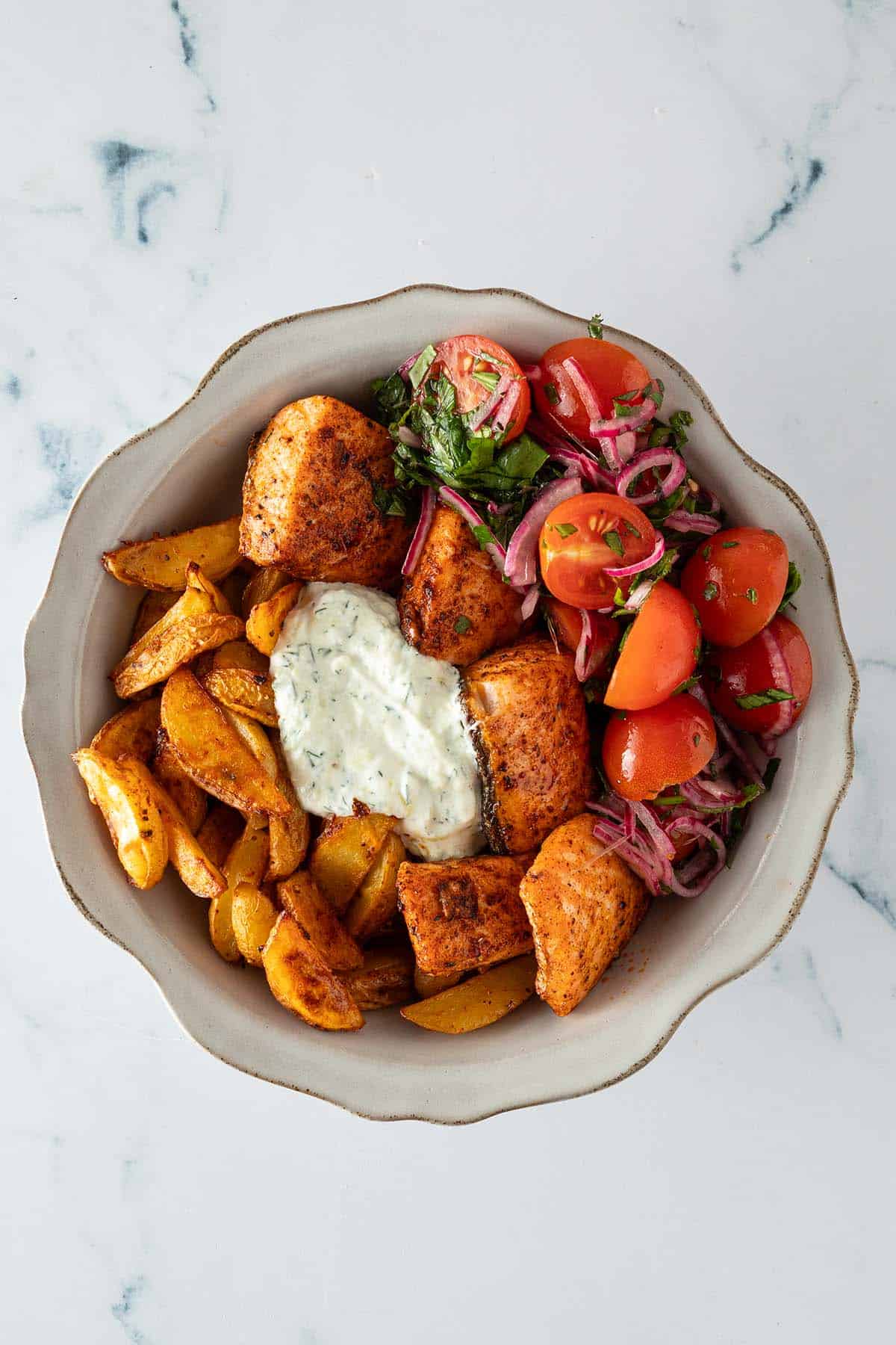 Fertig zusammengestellte Bowl aus knusprigen Kartoffelspalten, Lachs-W&uuml;rfeln, Tomatensalat und Tzatziki.
