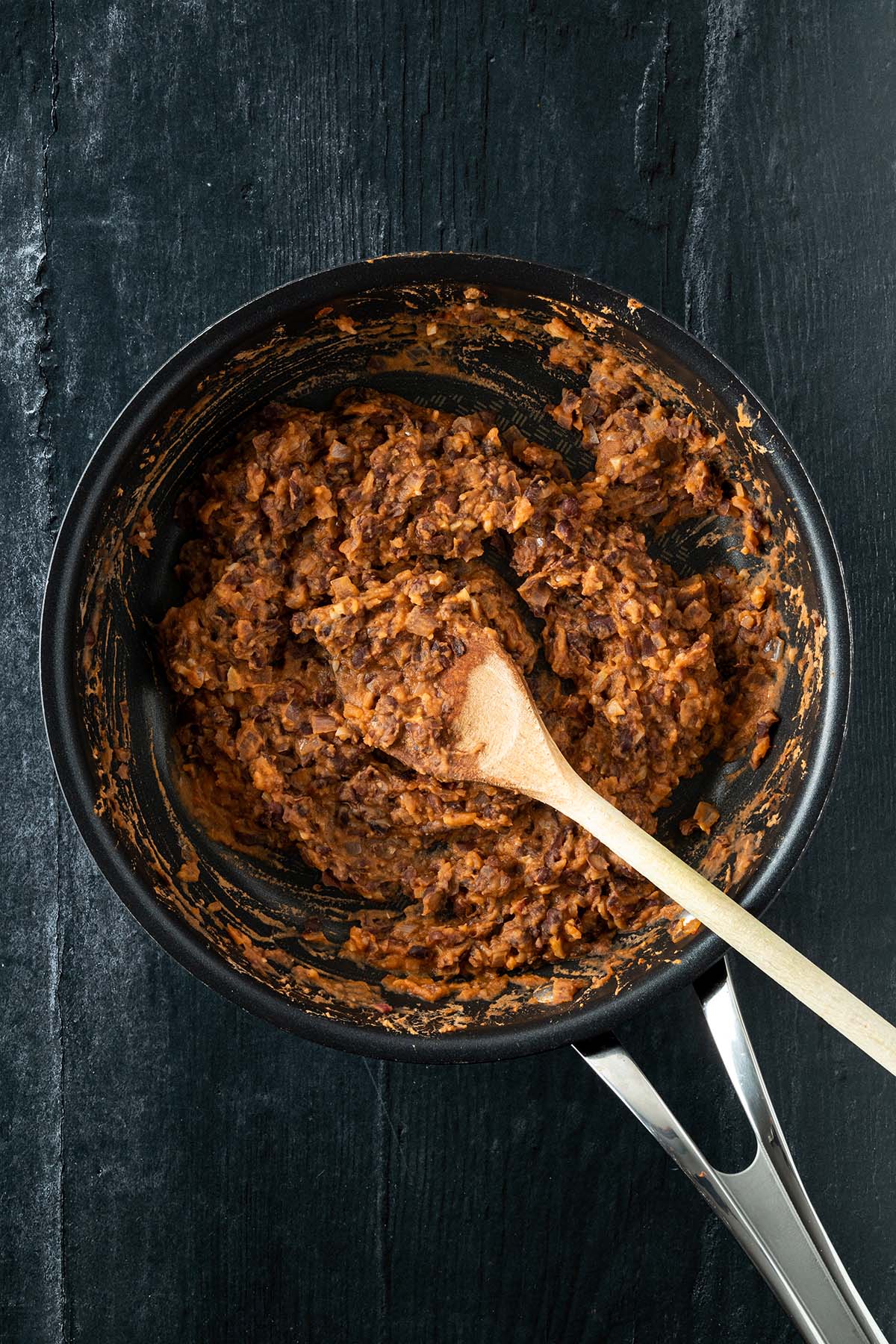 Vogelperspektive auf eine Pfanne mit Refried Beans auf einem dunklen Holzuntergrund.