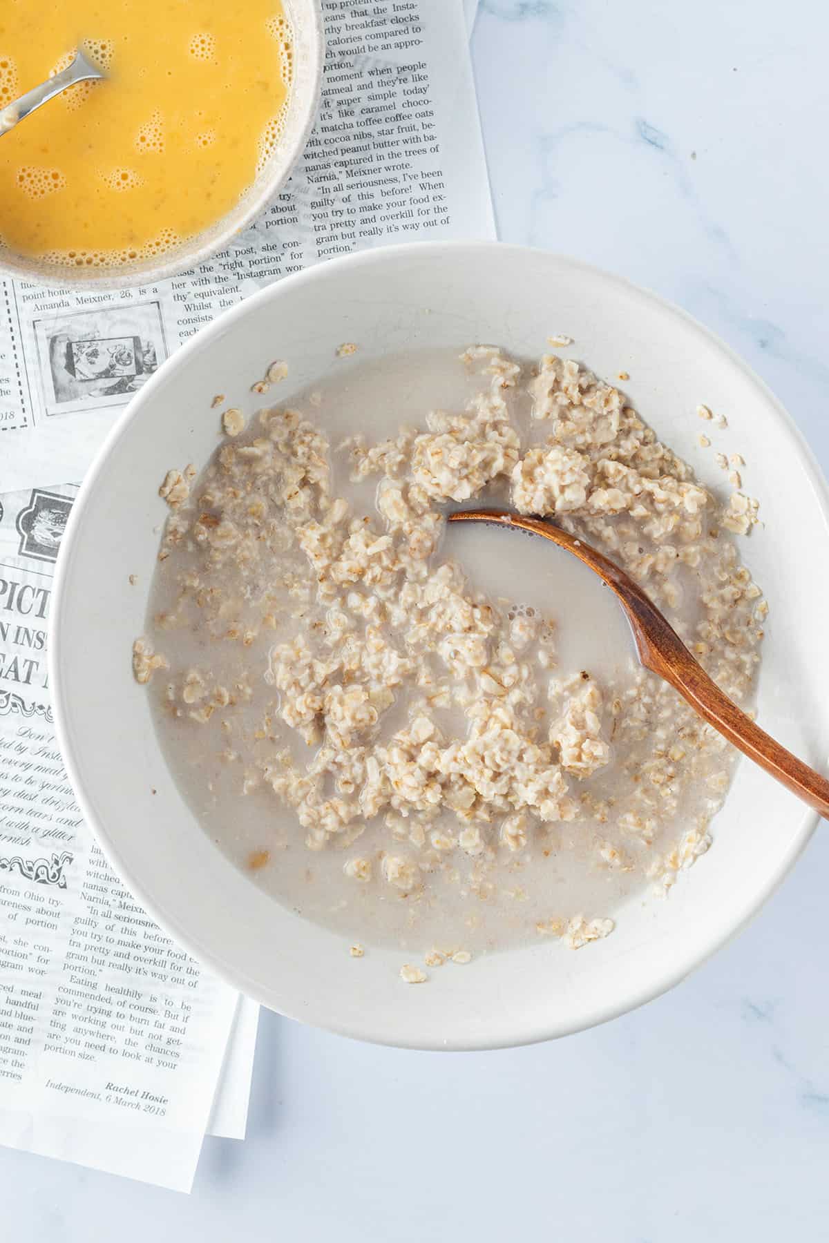 Eine Sch&uuml;ssel mit weichen Haferflocken und etwas Milch auf einem hellen Untergrund, daneben eine Sch&uuml;ssel mit verquirltem Ei.